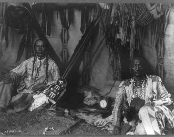 Image: Photograph of Little Plume and son Yellow Kidney sitting in a lodge. Taken by Edward Curtis in 1910. From the Library of Congress.